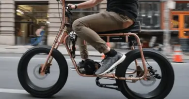 Side view of person, from the waist down, riding tan-coloured e-bike down a city street. The person is wearing a black t-shirt, grey pants, black socks and grey sneakers.