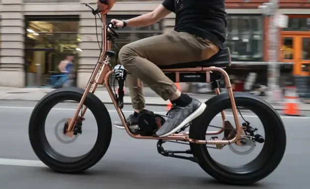 Side view of person, from the waist down, riding tan-coloured e-bike down a city street. The person is wearing a black t-shirt, grey pants, black socks and grey sneakers.