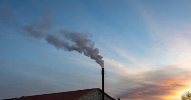 A close-up of a heating pipe on top of a house, releasing black smoke into the environment during a sunset.