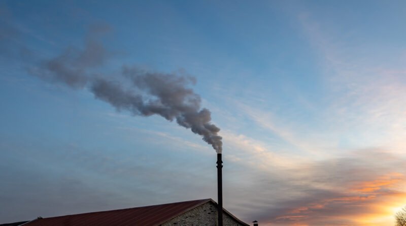 A close-up of a heating pipe on top of a house, releasing black smoke into the environment during a sunset.