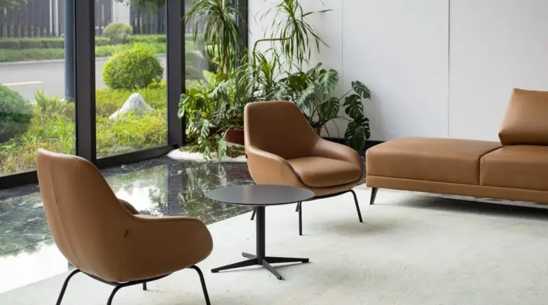 A brown leather sofa and two chairs inside a room with concrete floors, a small pond with plants, and large windows.