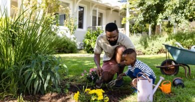 A man and a boy planting pink and yellow flowers in the home garden, next to a wheelbarrow and two water containers.