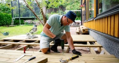 A man drills deck boards beside a yellow house while tools and a trampoline fill the yard behind him.