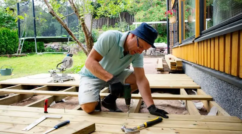 A man drills deck boards beside a yellow house while tools and a trampoline fill the yard behind him.
