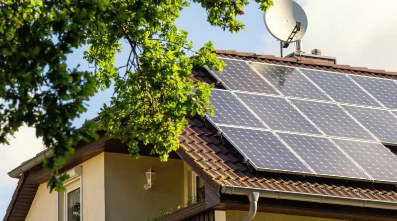 A two-story home with multiple rows of solar panels next to a large dish antenna, and a tree at the front of the house.