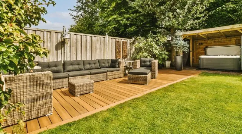 A backyard with a wooden fence, woven outdoor furniture with grey cushions, and a hot tub under a small pergola.