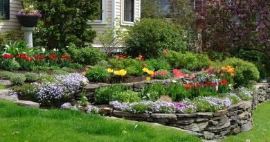 A stacked stone retaining wall borders a colorful flower garden as a green lawn slopes away from a beige house.