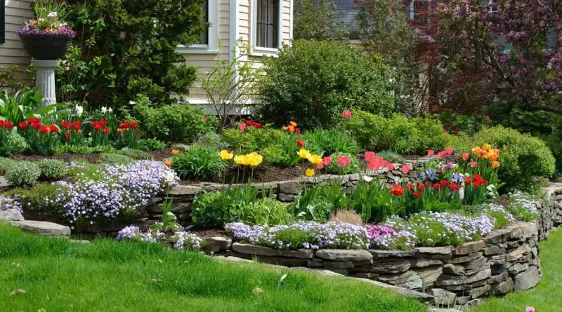 A stacked stone retaining wall borders a colorful flower garden as a green lawn slopes away from a beige house.