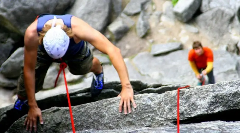 Rock climber wearing a helmet and harness scales a steep granite cliff using a red safety rope while a belayer assists from below.